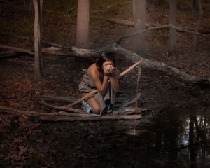 Enduring Hearts "Manitou Hill" by Jeremey Dennis. Photograph of a Native American in traditional clothing drinking from a stream.